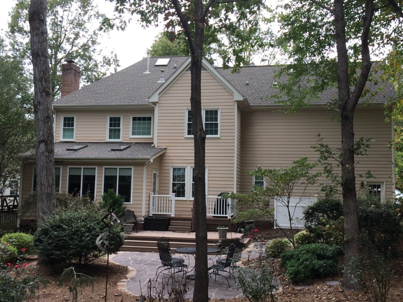A beige two-story house with Carolina siding and white-trimmed windows stands in Raleigh, NC, beside a shaded backyard patio.