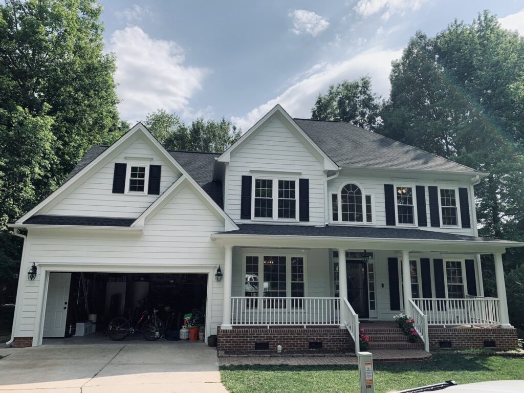 A Raleigh two-story white home with Carolina Hardie siding, black shutters, and double garage sits under a partly cloudy sky.