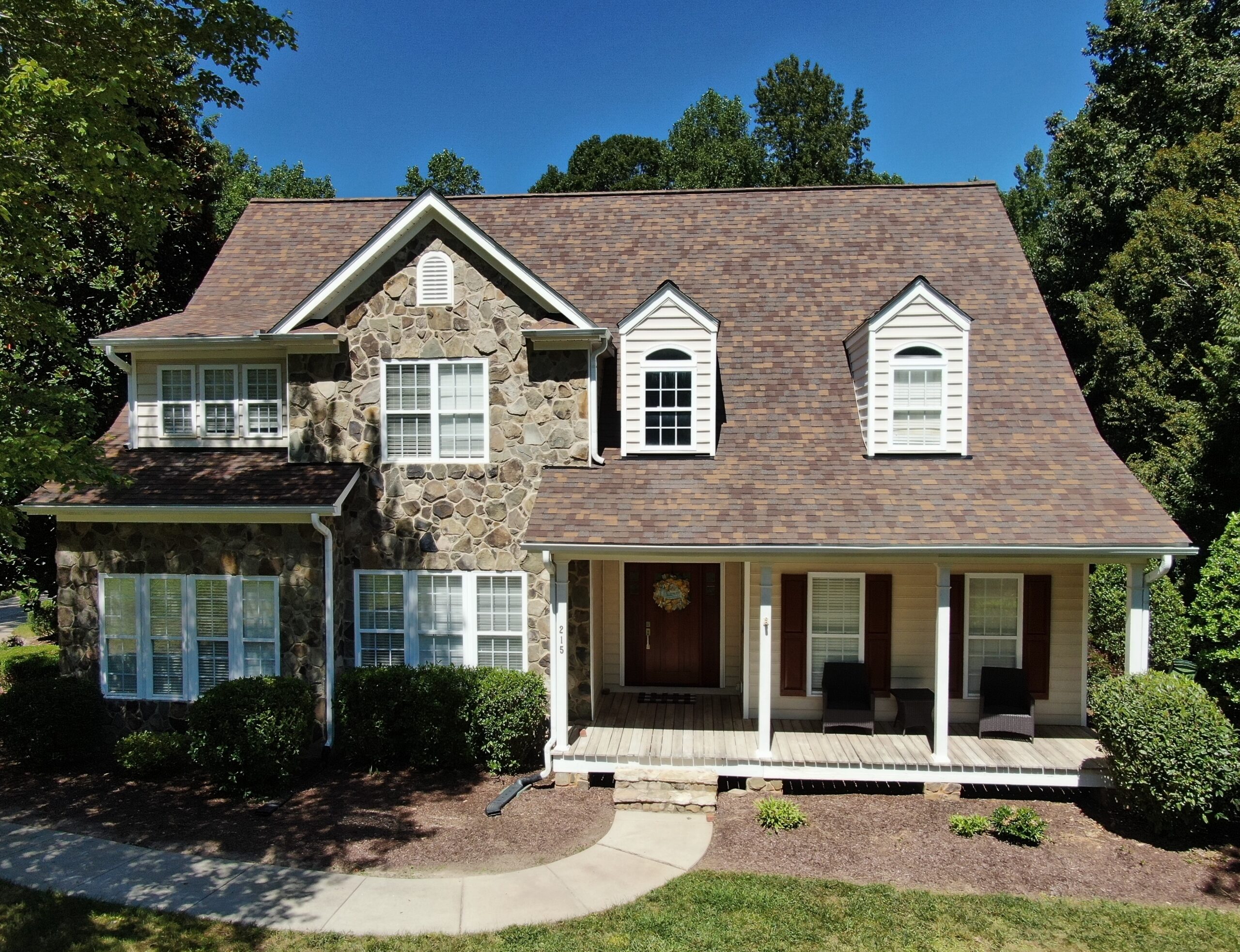 A two-story house with stone and beige siding, brown shingle roof by Carolina Roofing Raleigh/Apex, NC; white-columned porch.
