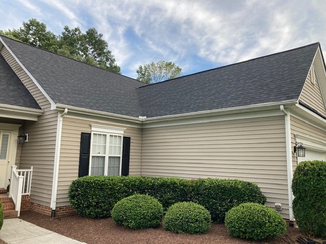 A beige house with white trim and black shutters, featuring a gabled roof by Carolina Roofing in Raleigh, NC, under a cloudy sky.