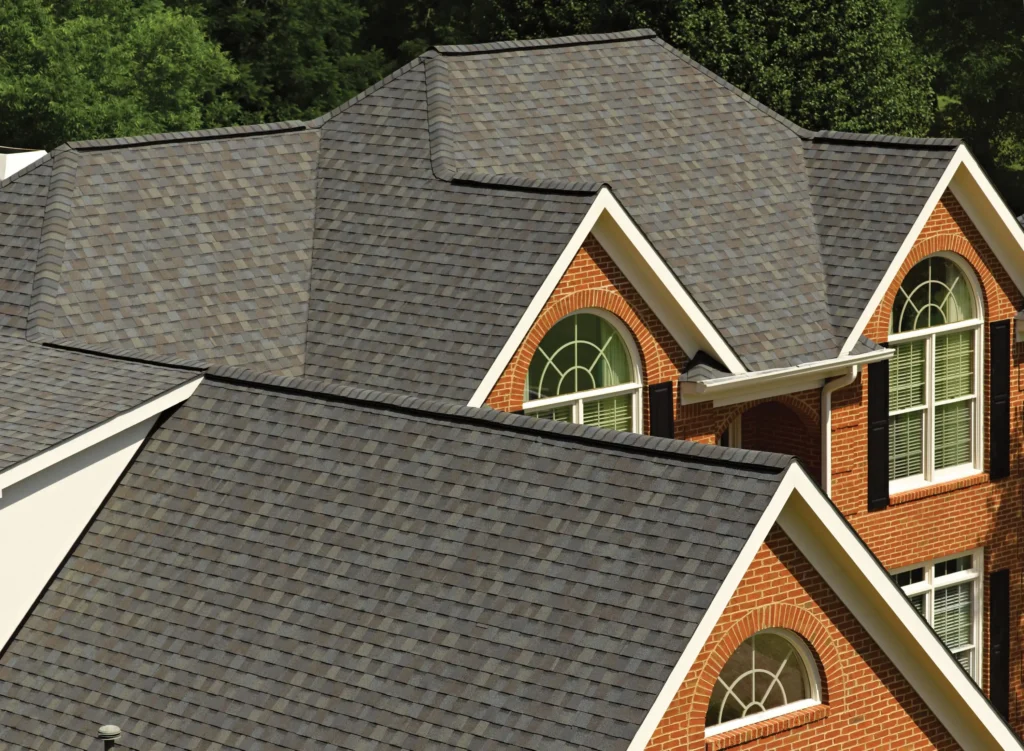 Close-up of gray asphalt shingle roof with gables and dormers by Carolina Roofing, Raleigh & Apex, NC; brick exterior, lush trees.