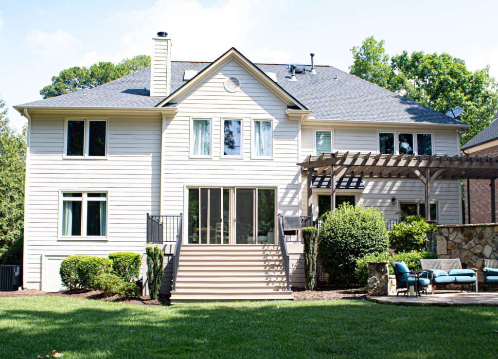 A two-story beige house in the Raleigh and Apex, NC area features newly installed Carolina siding, large energy-efficient windows, and a durable gray roof that highlights a recent update. Wooden stairs lead to a welcoming patio with a stylish pergola and comfortable outdoor furniture surrounded by neatly landscaped bushes. The backyard includes lush green grass, mature trees in the background, and a stone wall on the right, reflecting quality craftsmanship in local siding and roofing upgrades.