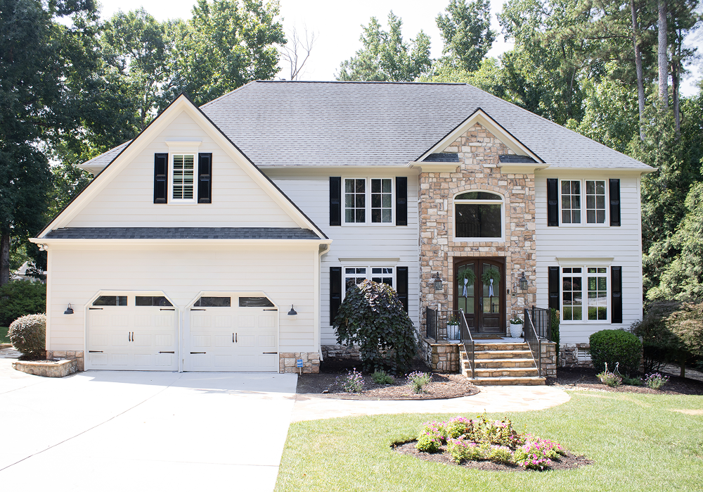 Two-story house featuring durable, attractive Carolina stone and white siding, accented by black shutters and a double garage. Spacious front windows showcase quality local installation, while the wooden double front doors lead up classic stone steps. The landscaped yard includes green grass, shrubs, flowers, and tall trees in the background—reflecting custom exterior work found in Raleigh and Apex, NC. Natural sunlight enhances the expertly crafted siding and windows for a bright transformation that highlights superior home improvement features tailored to the Carolinas.