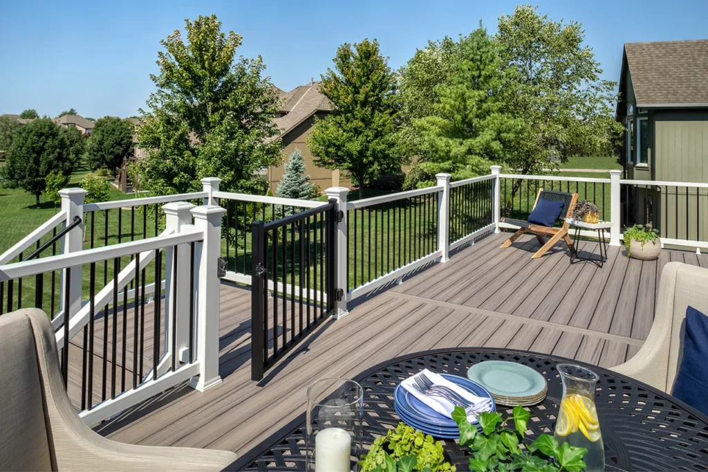 A spacious backyard Trex deck with gray railings overlooks a lush green lawn and mature trees, complementing well-maintained Carolina siding and roofing on nearby suburban homes in Raleigh. On the deck, two lounge chairs sit beside a black metal table set with plates, glasses, and a pitcher filled with lemon water—showcasing an inviting outdoor space to enjoy after upgrading your home’s exterior or windows in the Raleigh and Apex, NC area.
