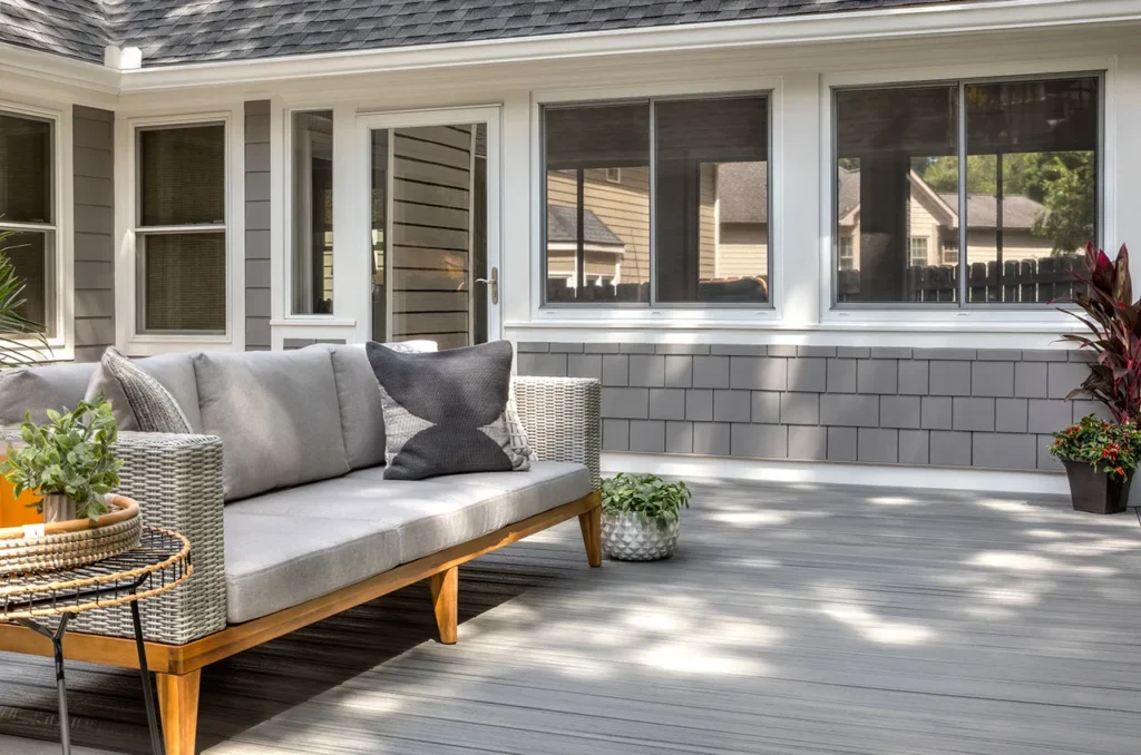 A modern outdoor Trex deck features light gray wood flooring complemented by a wicker sofa with gray cushions and throw pillows, alongside a small round side table topped with potted plants. Additional potted greenery accents the area near the house’s durable gray-shingled exterior and expansive energy-efficient windows—hallmarks of premier Carolina siding, window, and roofing installations in Raleigh and Apex, NC—making it an inviting space for relaxing afternoons while showcasing quality craftsmanship built to last.