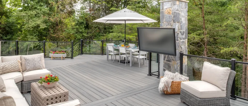 Spacious Trex deck with light gray flooring, cushioned chairs, wicker ottoman, flower pots, dining set under striped umbrella, stone pillar, large TV on stand, and glass railing—all surrounded by lush green trees. The Raleigh homeowner enjoyed a stylish outdoor retreat built quickly by Carolina Siding & Windows in Raleigh and Apex, NC—highlighting quality workmanship that ensures both lasting durability and impressive curb appeal.