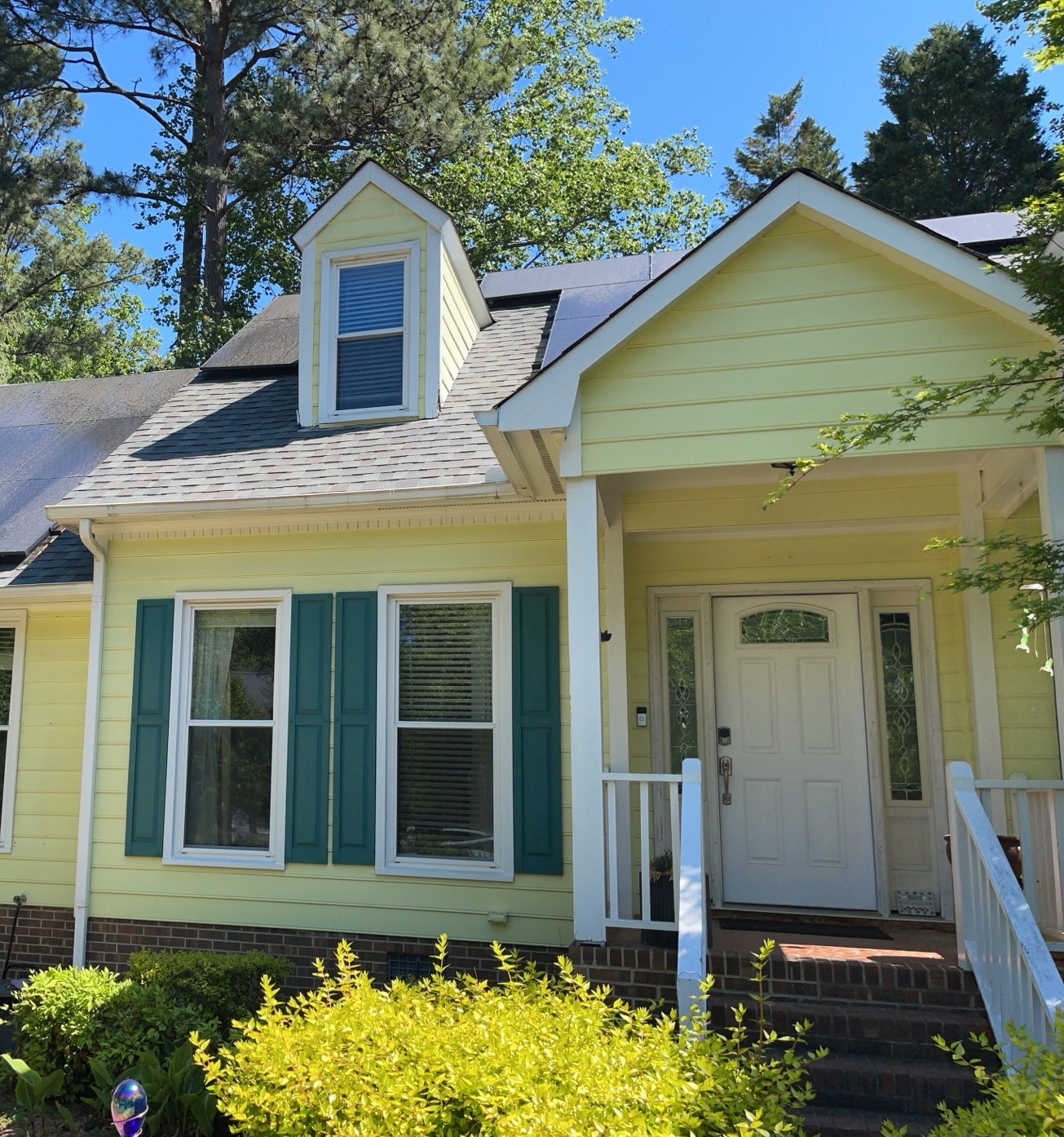A yellow house featuring Carolina siding with teal window shutters, a white front door, and a small porch with railing sits beneath a dormer window. The home is surrounded by green bushes and trees under a clear blue sky—an inviting example of residential roofing and windows in Raleigh and Apex, NC.