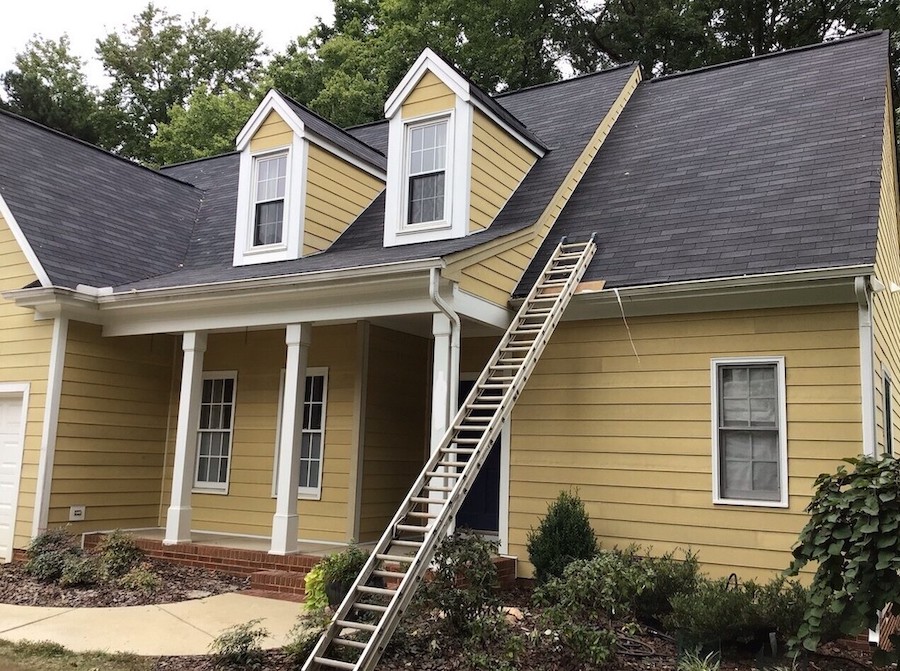 A yellow house with white trim features a steep, dark gray roof and two front-facing dormer windows, illustrating common home styles found in Raleigh and Apex, NC. A large wooden ladder leans from the ground to the roof, drawing attention to essential maintenance considerations for homeowners comparing siding options like hardie board or brick. The inviting porch is supported by four white columns, complemented by neatly trimmed bushes in front and mature trees in the background—an exterior well-suited for top-quality Carolina siding, window upgrades, or roofing services.