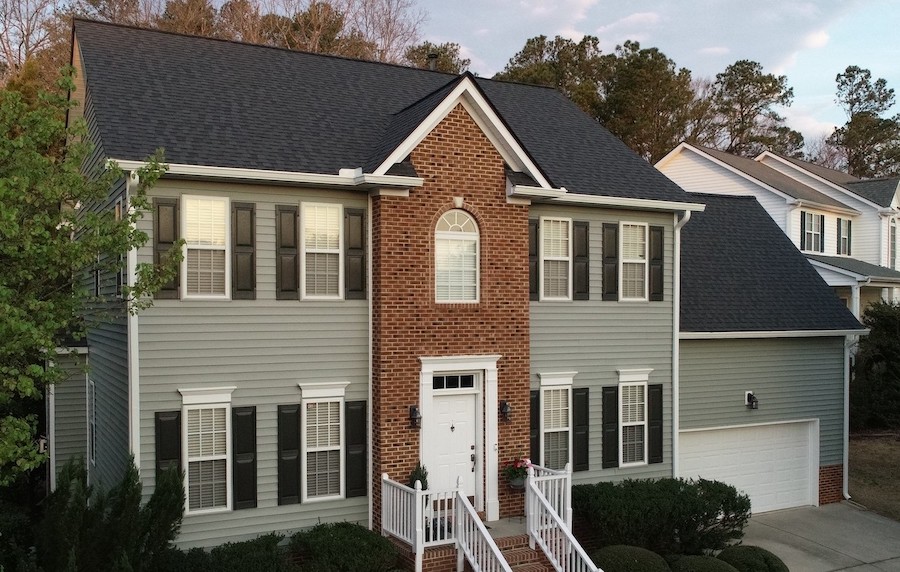 Two-story suburban home in Raleigh or Apex, NC, showcasing gray hardie board siding accented with brick around the central entrance, highlighted by white trim and black shutters beneath a dark roof. The inviting white front door features steps and railing, with neatly manicured bushes providing added curb appeal. An attached two-car garage is situated on the right, while mature trees create a picturesque backdrop—demonstrating why local homeowners choose this blend of durable hardie board siding and brick for both beauty and weather resistance. This exterior also illustrates the region’s preference for quality siding installation, energy-efficient windows, and reliable roofing solutions from Carolina specialists.