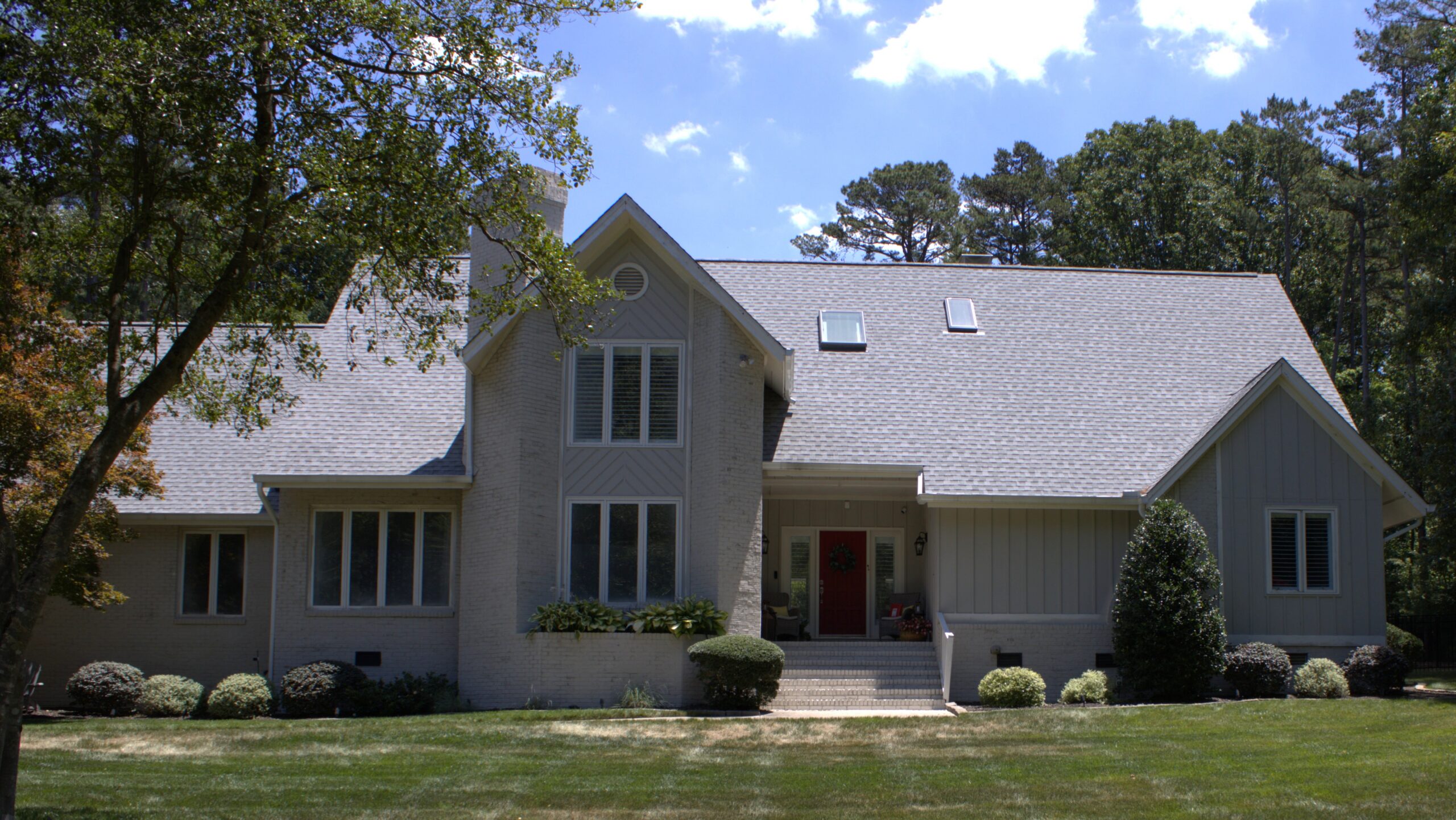 Two-story, light-gray house in Raleigh or Apex, NC, featuring Carolina-style siding and expansive windows that welcome natural light. The home has a steep gray roof with skylights installed by Carolina roofing specialists, and a welcoming front porch with a bold red door. Well-manicured bushes and lawn complement the home’s exterior, while tall trees frame the property against a backdrop of blue sky and soft clouds. Sunlight casts gentle shadows on the façade, highlighting quality local craftsmanship in siding, windows, and roofing.
