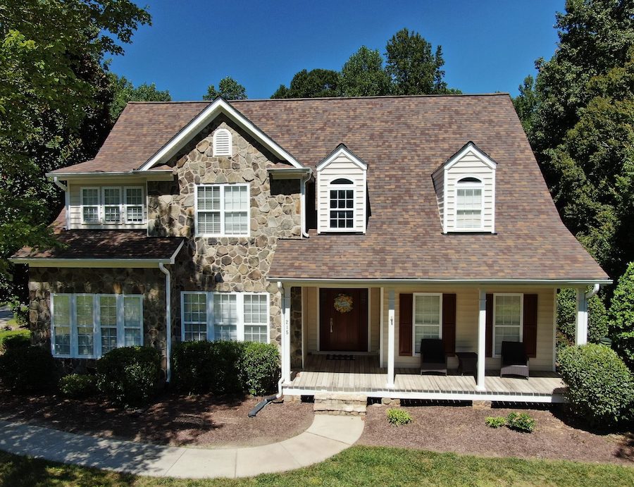 Two-story house in the Raleigh and Apex, NC area features a combination of stone, Hardie Board, and siding exterior for versatile curb appeal. The home showcases Carolina-style siding with a large gabled roof tailored for regional weather, three dormer windows that enhance natural light, energy-efficient windows and white trim. The wide front porch with columns offers southern charm, while manicured shrubs and a curved sidewalk add to the welcoming entrance—all surrounded by mature trees under a clear blue sky.