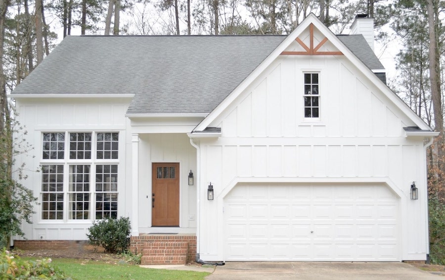 A modern white house in Raleigh or Apex, NC, features vertical siding by Carolina Siding & Windows, large windows on the left, a wooden front door with brick steps, two black lantern lights, a double garage door, and a triangular accent above a small window. The recently updated siding not only boosts curb appeal but can also raise your home’s value—perfect if you’re selling in the Triangle area. Surrounded by lush trees and greenery.