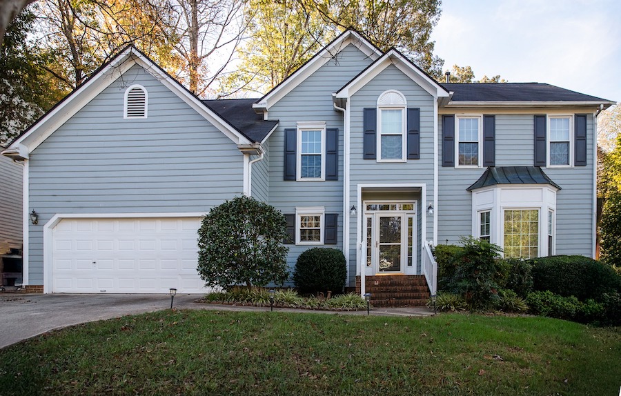 Two-story blue house with white trim and shutters, featuring Carolina siding that enhances durability and curb appeal. Double garage on the left, a welcoming front porch with white columns, steps leading to the door, and two large bushes by the entrance. A bay window on the right showcases energy-efficient Carolina windows ideal for Raleigh and Apex, NC homes. Well-maintained siding boosts home value in this area. Leafless trees in the background and a grass lawn in front complete the scene.