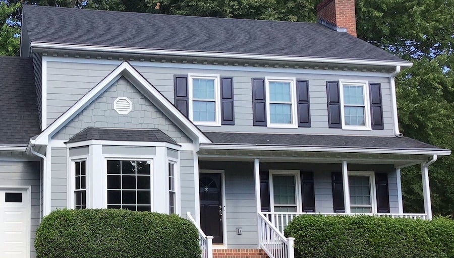 Two-story gray house with white trim, black shutters, a brick chimney, and a bay window on the left. There’s a small porch with white railings, steps, and hedges in front. The roof is dark gray, framed by tall green trees in the background. Recently updated siding from Carolina Siding in Raleigh and Apex, NC boosts curb appeal—an ideal upgrade for homeowners looking to increase property value or prepare their house for sale.