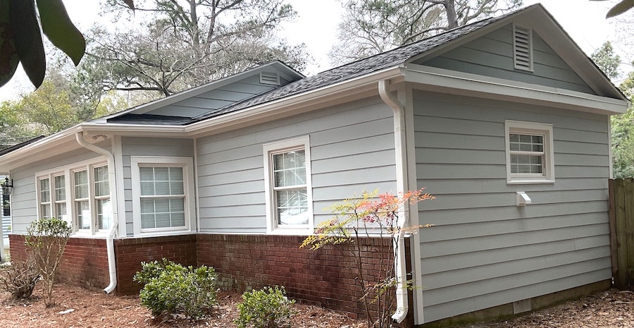 A single-story house in Raleigh or Apex, NC, features light gray horizontal siding and white trim installed by Carolina Siding experts, along with a sturdy brick foundation. Multiple white-framed windows provide modern curb appeal, complemented by well-maintained white gutters—all services offered by Carolina Siding & Windows. The home is surrounded by lush bushes and trees with pine straw spreading neatly across the yard, showcasing how professional siding, window, and gutter installations can significantly enhance your home’s value in the Triangle area.