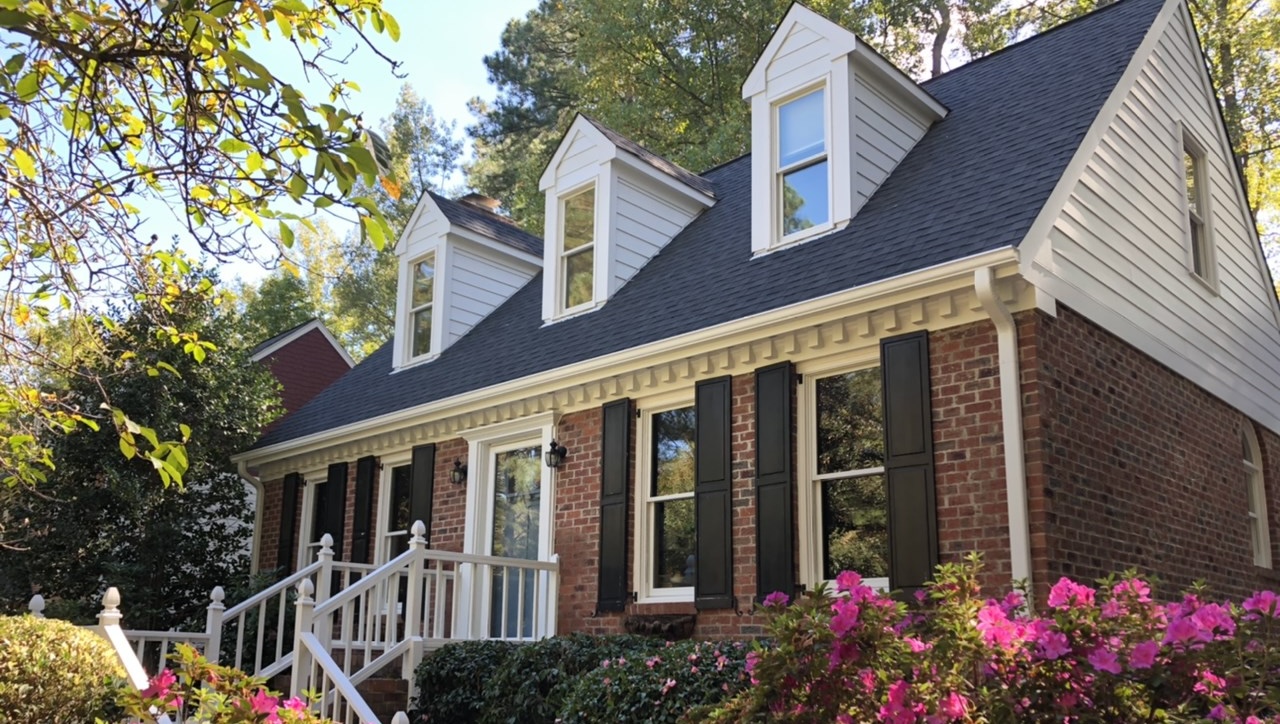 A red brick house with white Hardie siding on the upper level, a dark shingled roof, and three gabled dormers in Raleigh and Apex, NC. The exterior displays how to mix brick and Hardie siding with white trim, dark shutters, and a front railing, all surrounded by lush green trees and pink azaleas.